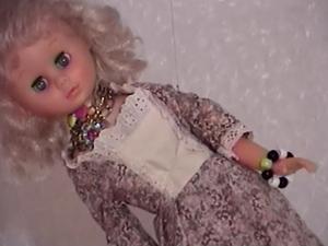 A feminine, kitsch style doll wearing tacky jewelry hangs from fishing wire in front of a bubble-wrapped background.